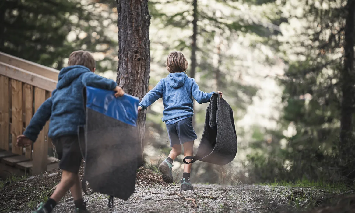 Zwei Kinder in blauen Jacken tragen Rutschmatten durch Lärchenwald am Speikboden bergauf