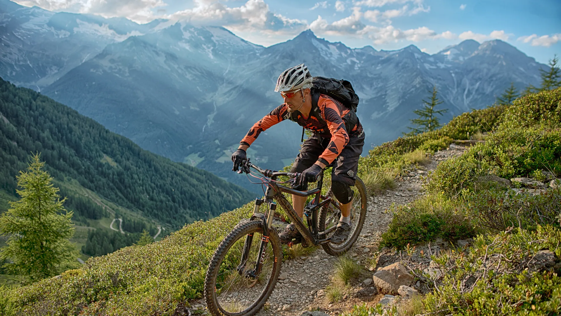 Mountain biker in orange gear riding a singletrack through alpine meadows with Zillertal Alps panorama
