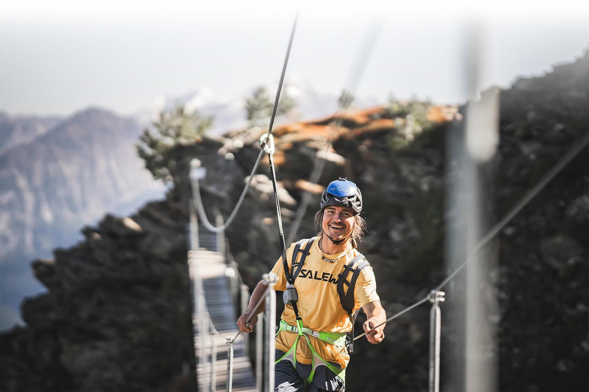 Laughing via ferrata hiker with blue helmet on suspension bridge, Ahrntal mountain panorama