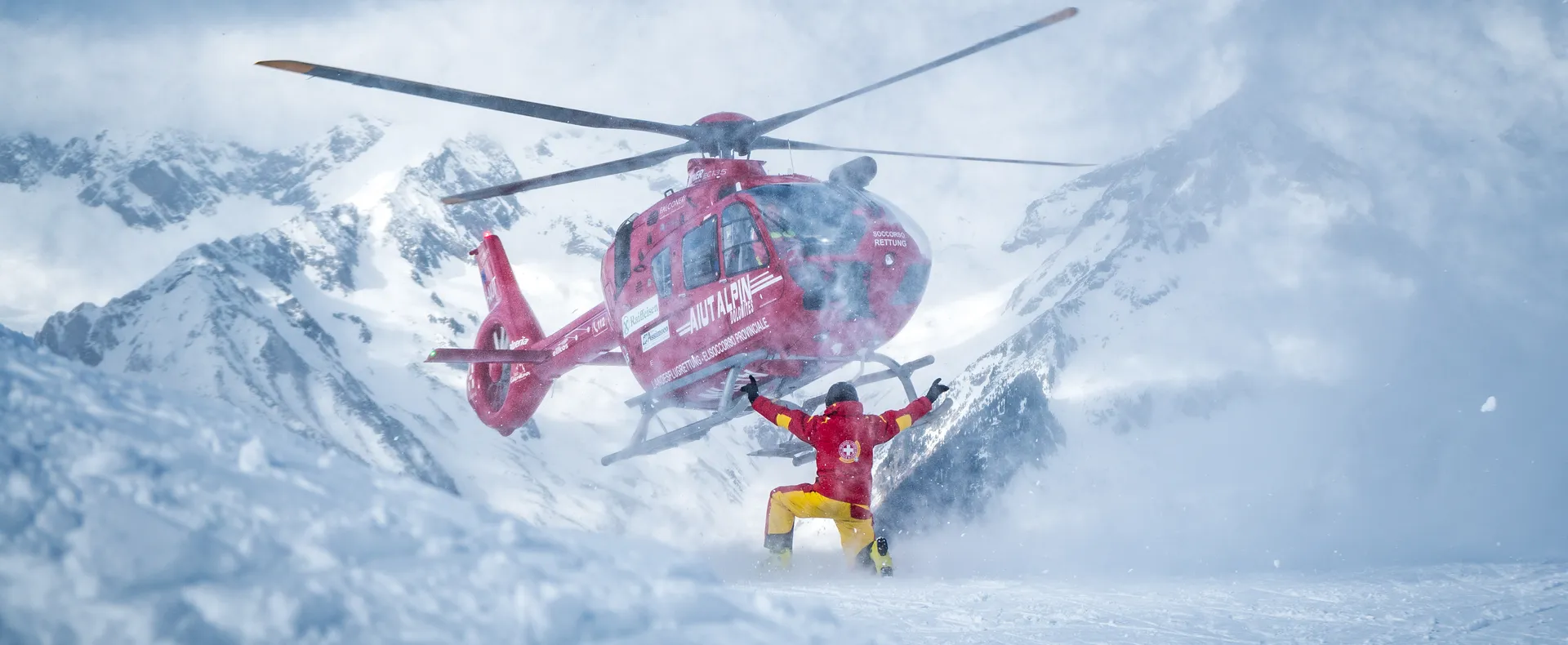 Red rescue helicopter Aiut Alpin Dolomites landing in deep snow during slope rescue operation
