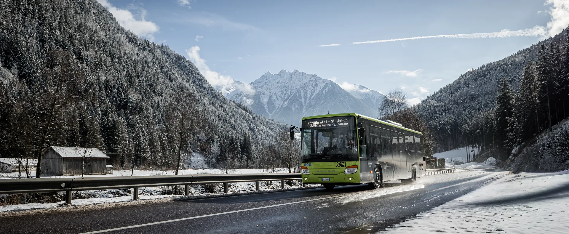 Grüner Skibus der Linie Ahrntal fährt durch verschneites Tal mit Nadelwald und Bergen