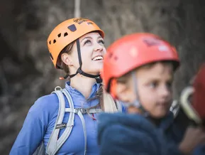 Young woman with orange Speikboden climbing helmet smiles radiantly, child with red helmet in front