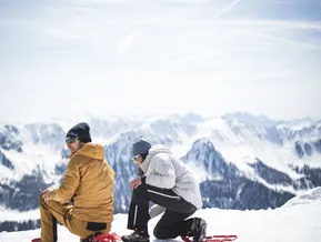 Coppia allaccia le ciaspolette rosse sul pianoro di vetta con vista sulle Alpi della Zillertal al Klausberg e Speikboden