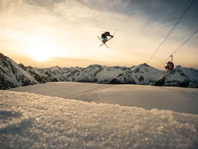 Freestyle-Skifahrer beim Cross-Trick mit roten Klausberg-Gondeln und Südtiroler Alpenpanorama