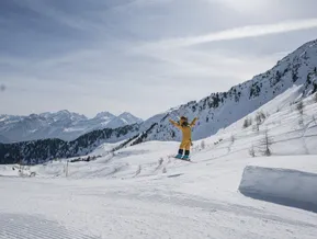 Das Skigebiet-Maskottchen im gelben Kostüm springt auf Skiern über einen Kicker im Ahrntal