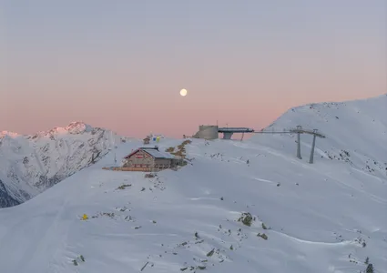 Bergstation mit Sessellift bei Sonnenuntergang und Vollmond über verschneiten Gipfeln