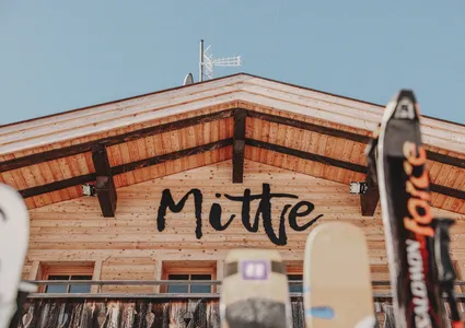 Hüttenschild mit Schriftzug Mitte und Skiern im Vordergrund, blauer Himmel