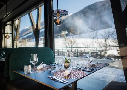 Fine-Dining Restaurant mit Panoramablick auf verschneite Berge und Sessellift, gedeckter Tisch mit Weinkelchen