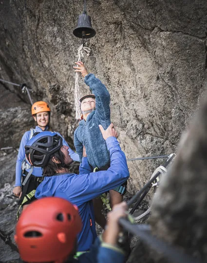 Familie am Klettersteig Ahrntal: Kind läutet Gipfelglocke, Eltern mit Helmen freuen sich