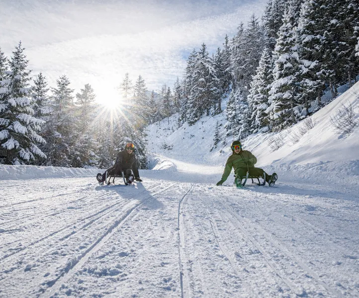 Due persone slittano al sole attraverso il bosco invernale innevato sulla pista da slittino in Val Aurina