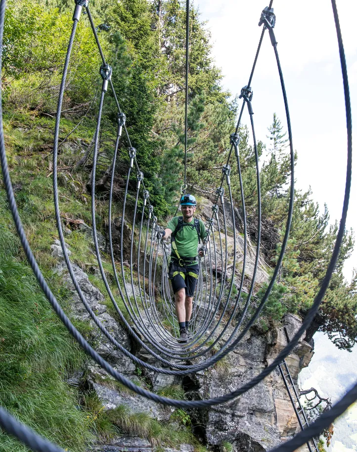 Climber crosses steel rope suspension bridge through Swiss stone pine forest, Ahrntal via ferrata