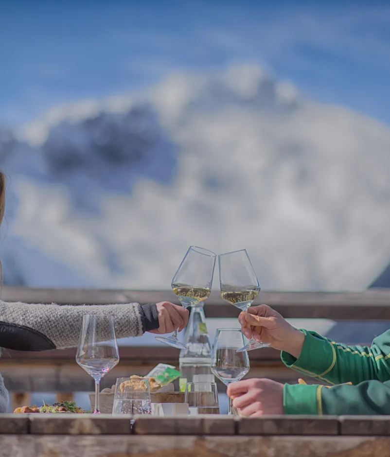 Couple toasting with white wine on a mountain hut terrace, snowy peaks in the background