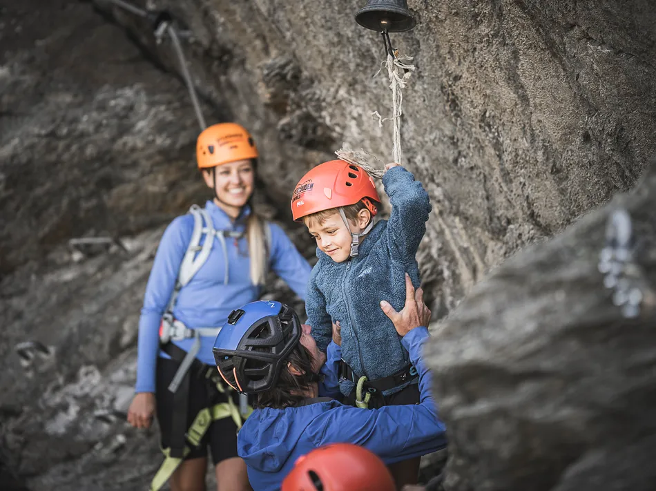 Family at Ahrntal via ferrata: father lifts son with helmet to summit bell, mother smiles along