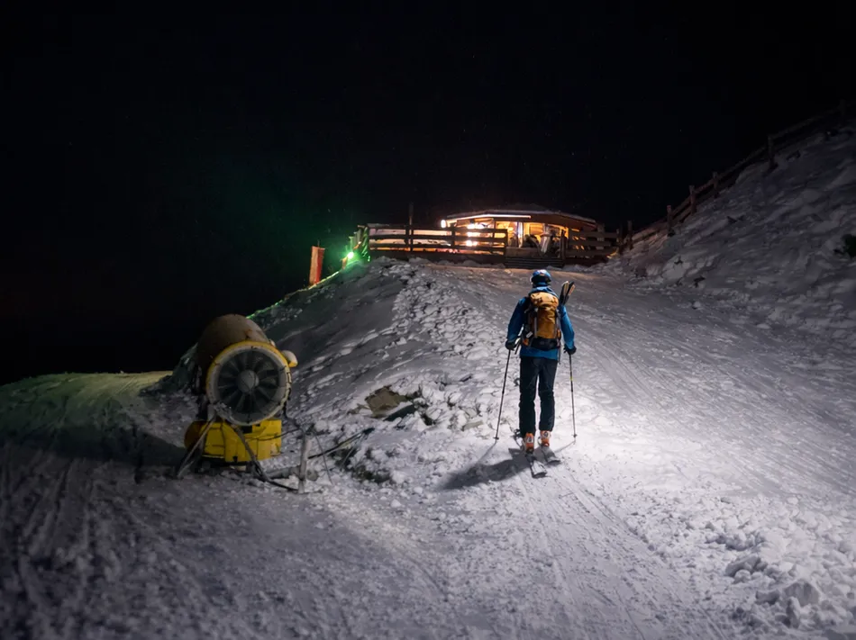 Ski tourer with headlamp ascending at night towards the illuminated mountain hut at Klausberg