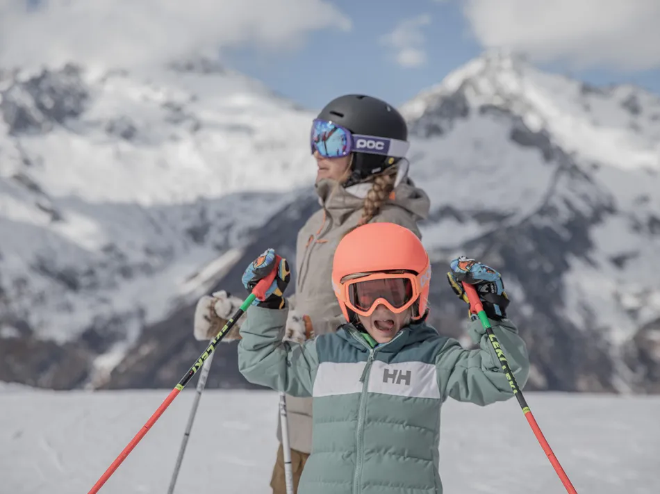 Cheering child with orange ski helmet raises poles on the slope, mother in the background