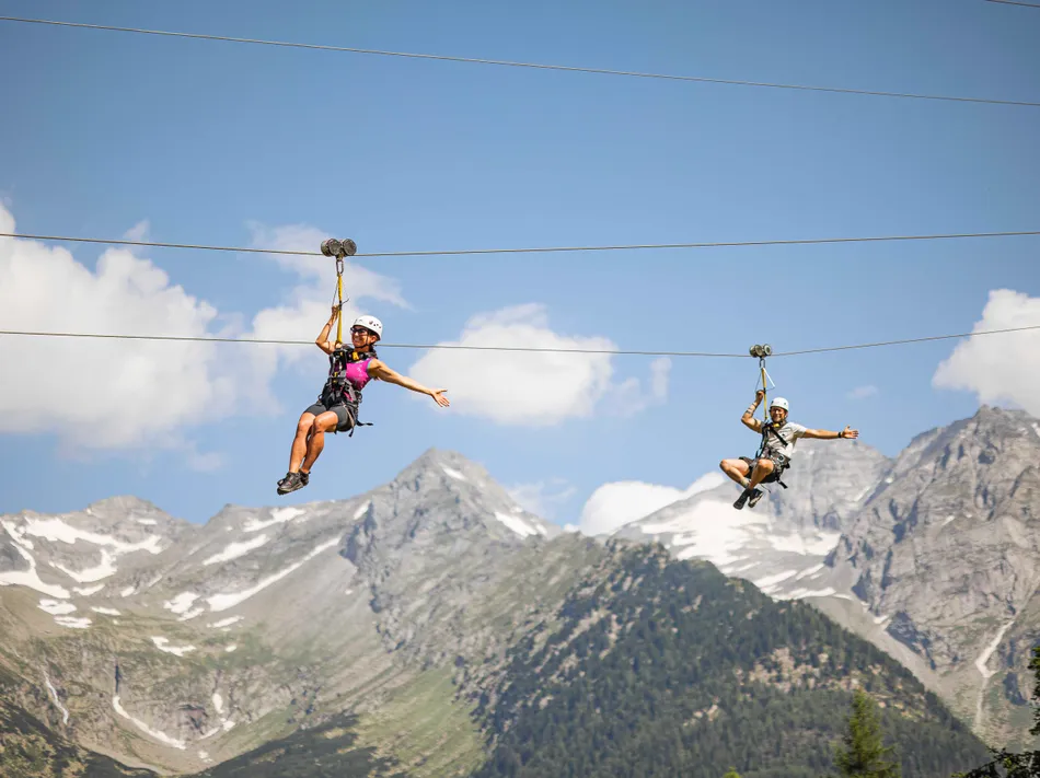 Two people zip along parallel ziplines through the air, Zillertal Alps in the background