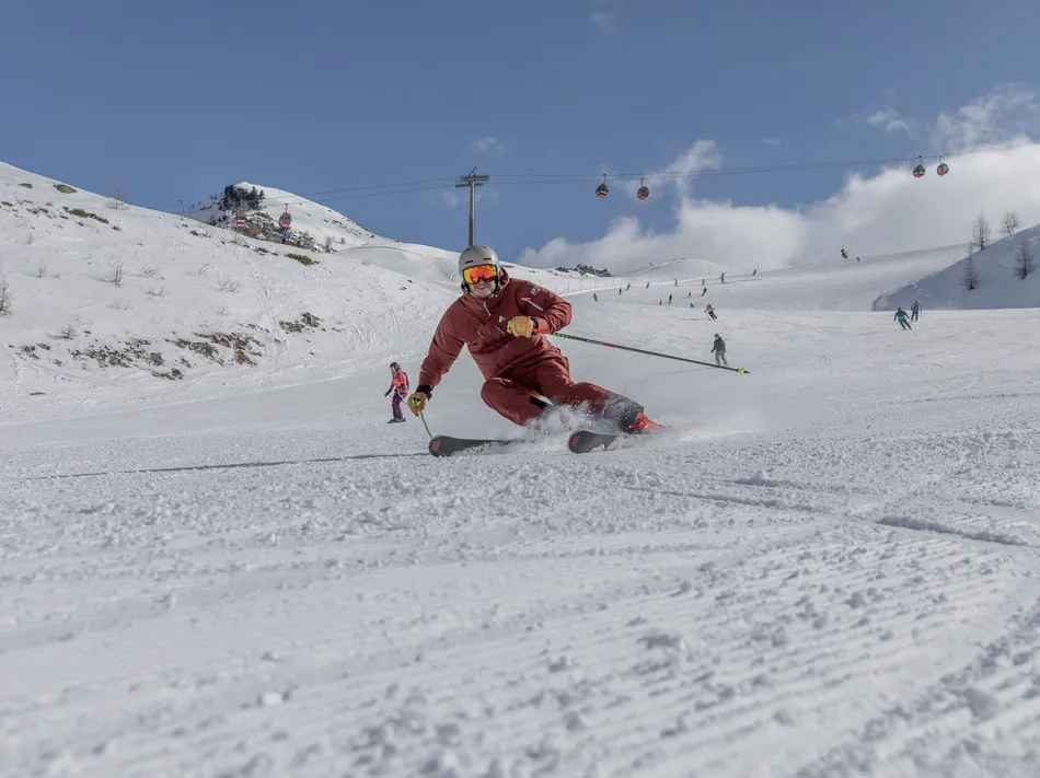 Skier in red suit carving on groomed slope with cable car in the background